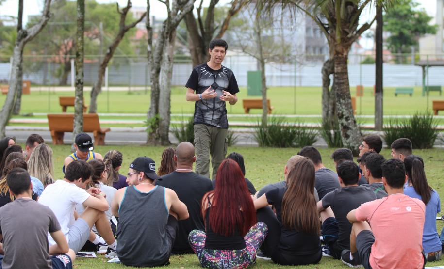 Professor em pé repassando orientações para acadêmicos que estão sentados na grama.